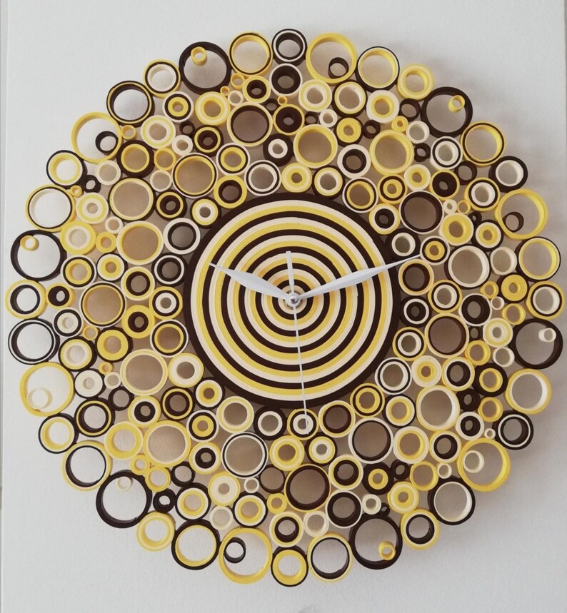 May include: A yellow, brown, and white paper quilling clock with a circular design. The clock face is made of concentric circles of yellow and brown paper. The clock hands are white.