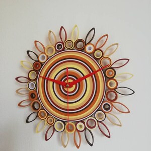May include: A circular clock with a sunburst design made of rolled paper. The clock face is a series of concentric circles in shades of brown, yellow, and white. The clock hands are red.