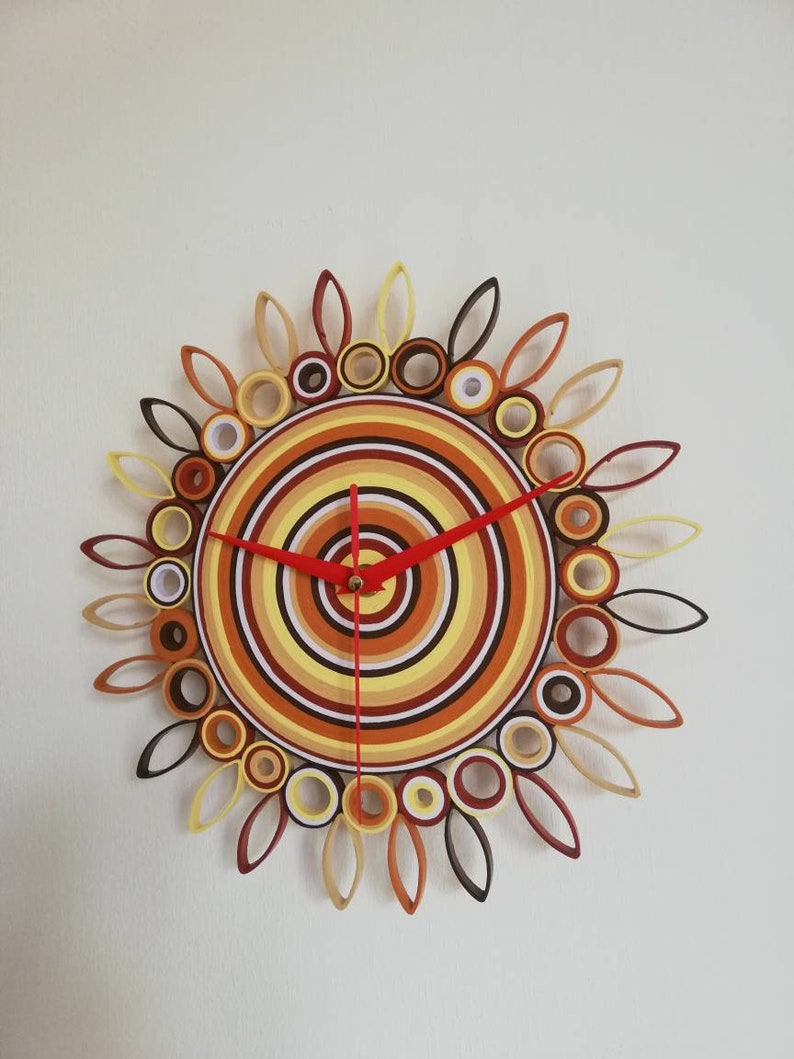 May include: A circular clock with a sunburst design made of rolled paper. The clock face is a series of concentric circles in shades of brown, yellow, and white. The clock hands are red.