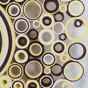 May include: A close-up of a wall art piece made of paper circles. The circles are arranged in a circular pattern and are painted in shades of yellow, white, and brown.
