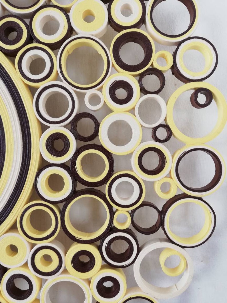 May include: A close-up of a wall art piece made of paper circles. The circles are arranged in a circular pattern and are painted in shades of yellow, white, and brown.