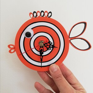 Op de afbeelding: Een grillige oranje en witte visvormige wandklok. De wijzerplaat heeft een doelontwerp met een wiebeloog. De klok heeft zwarte wijzers en decoratieve vinnen. De klok wordt door een hand vastgehouden.