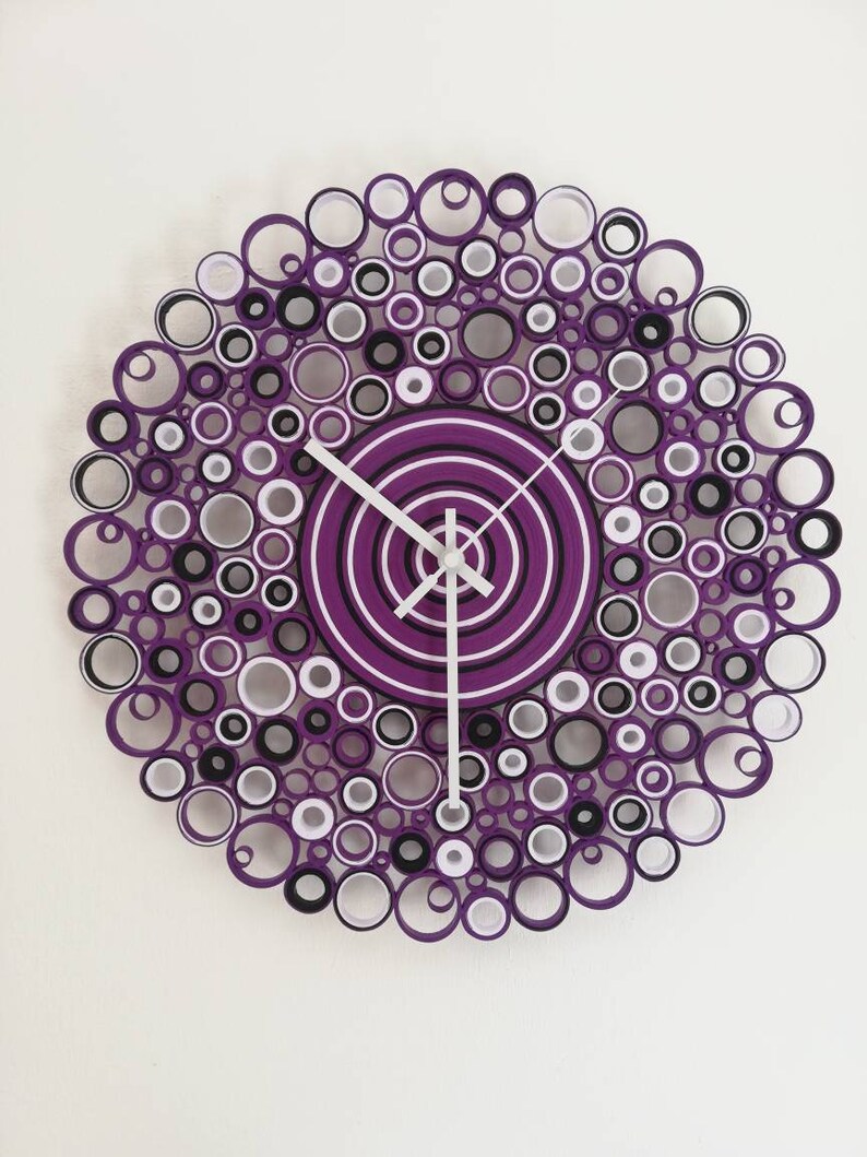 orologio da parete creato con rotolini di cartoncini colorati. i colori sono: viola predominante, bianco e nero. i rotolini hanno diversi diametri e sono incollati tra di loro, le lancette sono bianche e lunghe e il meccanismo è silenzioso