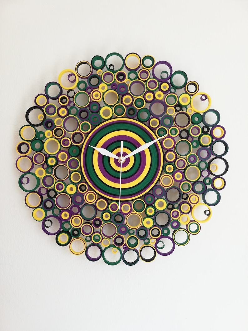 May include: A circular wall clock made of paper rolls, with a concentric circle design in green, yellow, and purple. The clock face is surrounded by a ring of smaller circles in black, green, yellow, and purple.