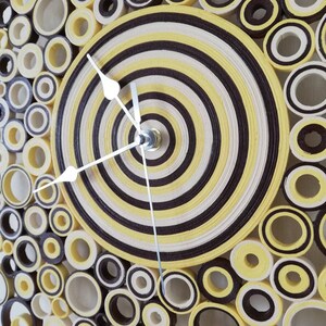 May include: A yellow and brown paper clock with a circular design. The clock face is made of concentric circles of yellow and brown paper. The clock hands are silver.