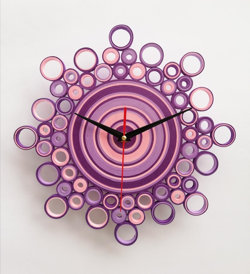 Orologio artigianale realizzato con la tecnica del quilling, composto da cerchi concentrici di carta nei toni del viola, glicine e del rosa. Il design ricorda un motivo floreale o una forma a raggiera, con un centro solido circondato da anelli di diverse dimensioni. Le lancette nere segnano l'ora. l'orologio è appeso ad un muro bianco