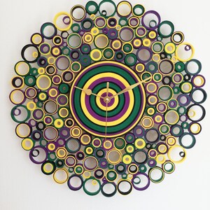 May include: A circular wall clock with a concentric circle design in shades of green, yellow, and purple. The clock face is surrounded by a ring of smaller circles in the same colors.