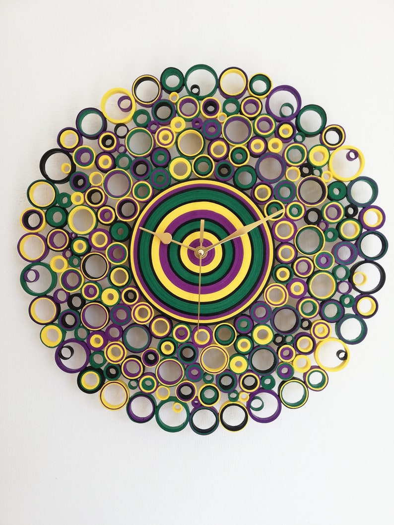 May include: A circular wall clock with a concentric circle design in shades of green, yellow, and purple. The clock face is surrounded by a ring of smaller circles in the same colors.