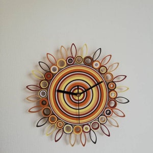 May include: A circular wall clock with a sunburst design. The clock face is made of paper and features concentric circles in shades of orange, yellow, and brown. The clock hands are black.