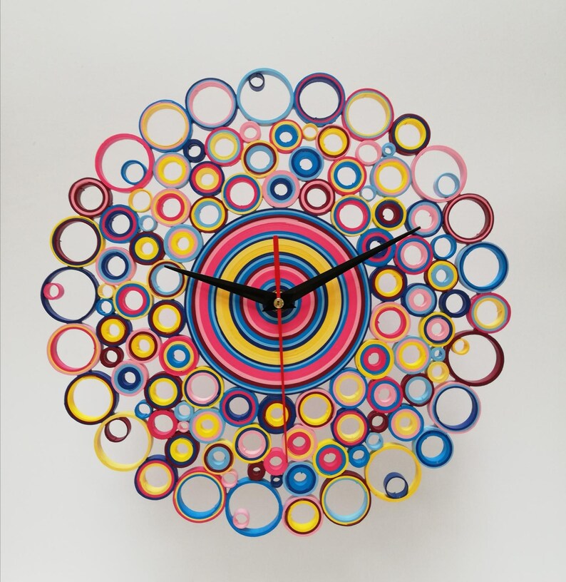 May include: A colorful clock made of paper rolls, with a red, yellow, and blue center and a black clock face. The clock is surrounded by a ring of smaller paper rolls in various colors, including pink, blue, yellow, and purple.