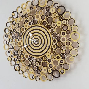 May include: A circular wall clock made of paper rolls in shades of yellow, brown, and cream. The clock face is a concentric circle design with a silver clock hand.
