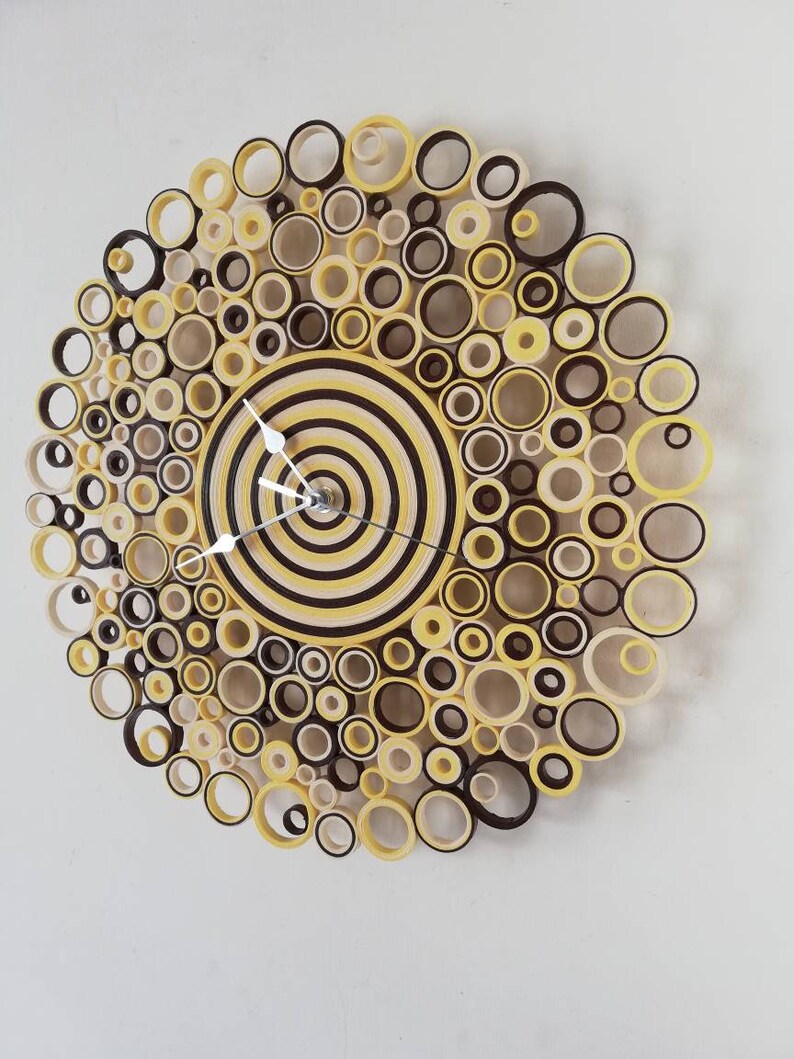 May include: A circular wall clock made of paper rolls in shades of yellow, brown, and cream. The clock face is a concentric circle design with a silver clock hand.