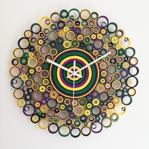 May include: A circular wall clock made of paper rolls, with a concentric circle design in green, yellow, and purple. The clock hands are white.