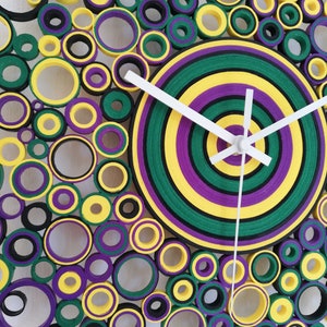 May include: A clock with a circular design made of layered paper circles in shades of green, yellow, purple, and black. The clock face is a concentric circle design with white hands.