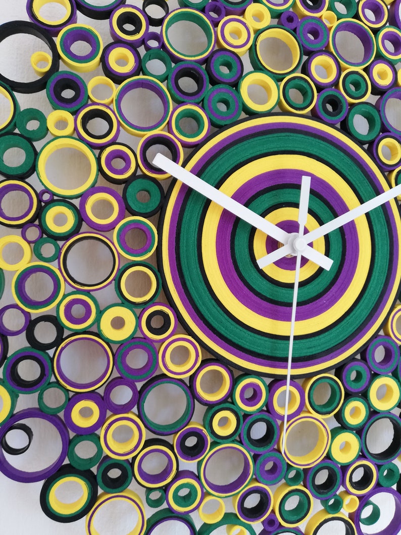 May include: A clock with a circular design made of layered paper circles in shades of green, yellow, purple, and black. The clock face is a concentric circle design with white hands.