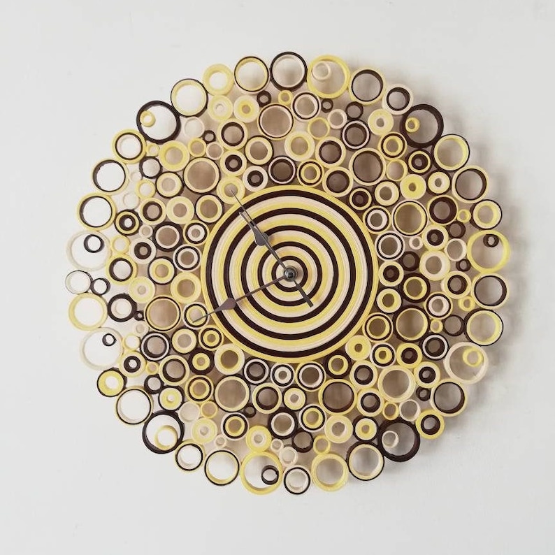 May include: A yellow and brown paper clock with a circular design. The clock face is made of concentric circles of yellow and brown paper. The clock hands are silver.