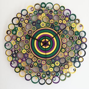 May include: A circular wall clock made of paper rolls, with a concentric circle design in yellow, green, and purple. The clock face is surrounded by a ring of smaller circles in black, yellow, green, and purple.