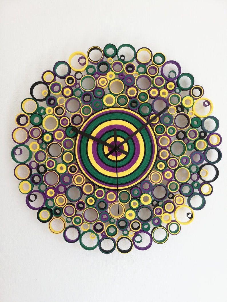 May include: A circular wall clock made of paper rolls, with a concentric circle design in yellow, green, and purple. The clock face is surrounded by a ring of smaller circles in black, yellow, green, and purple.