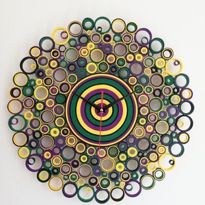 May include: A circular wall clock made of paper rolls, with a layered design of yellow, green, purple, and black circles. The clock face is a concentric circle design with black hands.