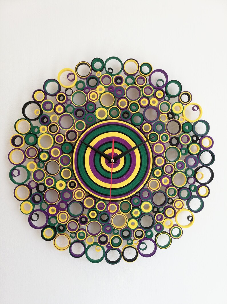 May include: A circular wall clock made of paper rolls, with a layered design of yellow, green, purple, and black circles. The clock face is a concentric circle design with black hands.