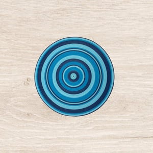 Puede incluir: Un posavasos redondo azul con círculos concéntricos en diferentes tonos de azul. El posavasos está centrado en una superficie clara con vetas de madera, creando un fondo contrastante. El diseño presenta un anillo exterior azul oscuro, con tonos más claros.