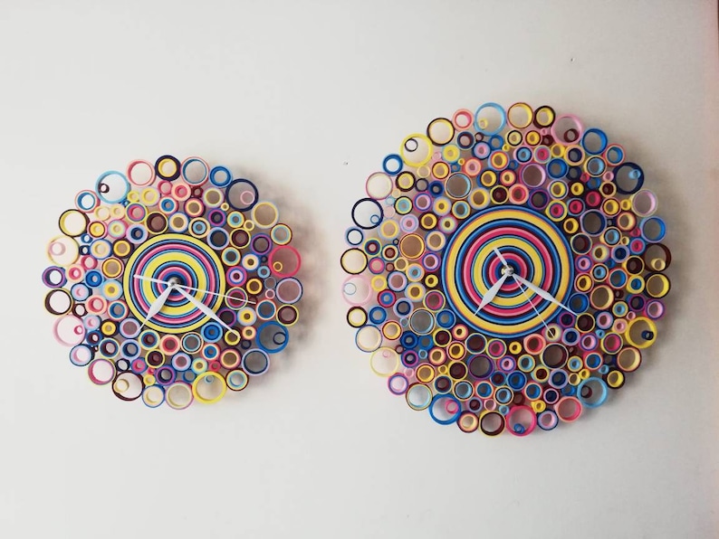 May include: Two colorful clocks made of paper circles, one larger than the other, with white hands and a concentric circle design in the center.