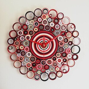 May include: A red, white, and black paper clock with a circular design. The clock face is made of concentric circles of paper, and the hands are red.