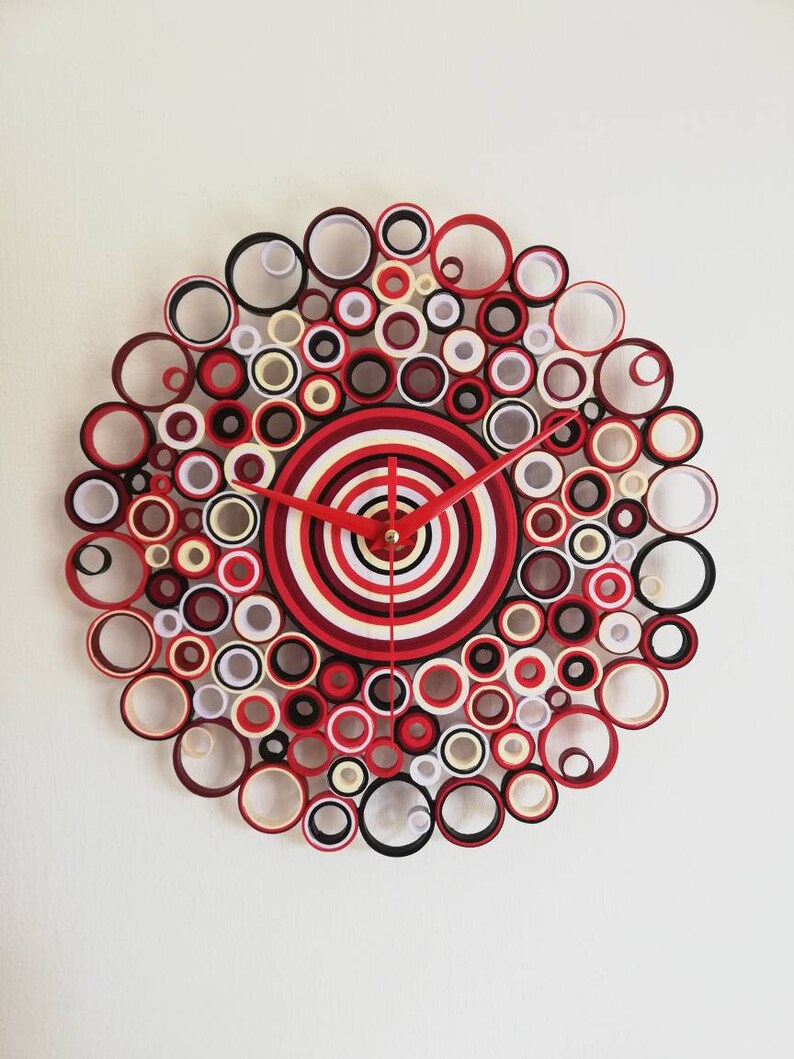 May include: A red, white, and black paper clock with a circular design. The clock face is made of concentric circles of paper, and the hands are red.