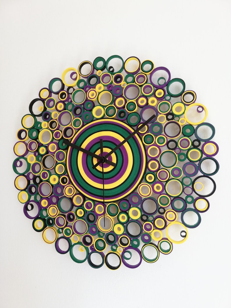 May include: A circular wall clock made of paper rolls, with a layered center in shades of green, yellow, and purple. The clock face is surrounded by a ring of smaller circles in black, yellow, green, and purple.