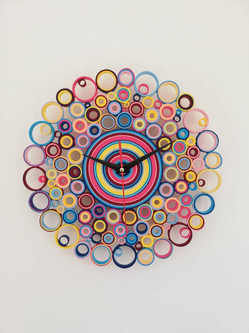May include: A colorful clock made of rolled paper circles in shades of pink, yellow, blue, and red. The clock face is a concentric circle design with black hands.