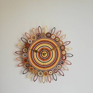 May include: A circular wall clock with a sunburst design made of paper. The clock face is a series of concentric circles in shades of orange, yellow, and brown. The clock hands are gold.