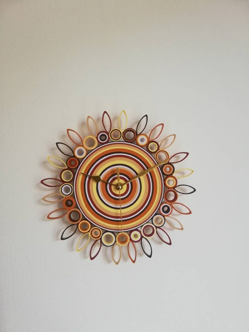 May include: A circular wall clock with a sunburst design made of paper. The clock face is a series of concentric circles in shades of orange, yellow, and brown. The clock hands are gold.
