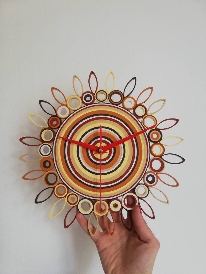 May include: A circular wall clock with a red, white, and brown concentric circle design. The clock has a red hour hand and a black minute hand. The clock is surrounded by a ring of brown, yellow, and black paper rolls.