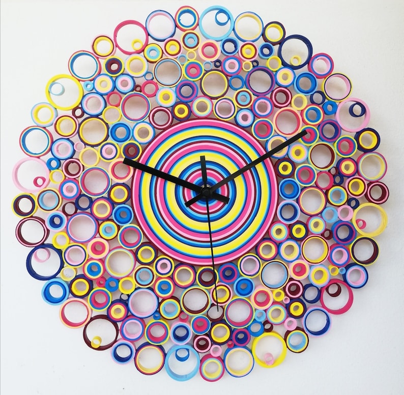 May include: A colorful clock made of paper circles, with a black clock face and hands. The clock is surrounded by a ring of smaller circles in various colors, including pink, blue, yellow, and green.