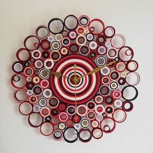 May include: A red, white, and black paper clock with a circular design. The clock face is made of concentric circles of paper, and the outer edge is decorated with a ring of smaller circles.
