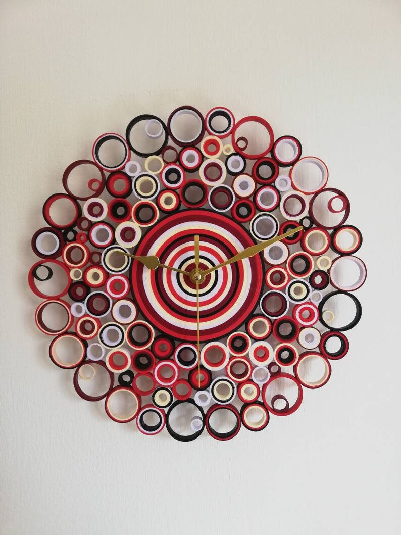 May include: A red, white, and black paper clock with a circular design. The clock face is made of concentric circles of paper, and the outer edge is decorated with a ring of smaller circles.
