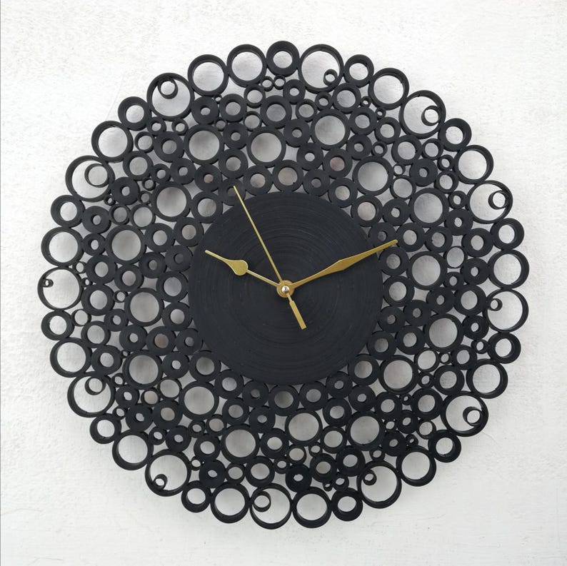 Un orologio da parete nero dal design unico con lancette dorate. La cornice è composta da cerchi neri di varie dimensioni, creando un motivo circolare decorativo. L'orologio è montato su una parete bianca.