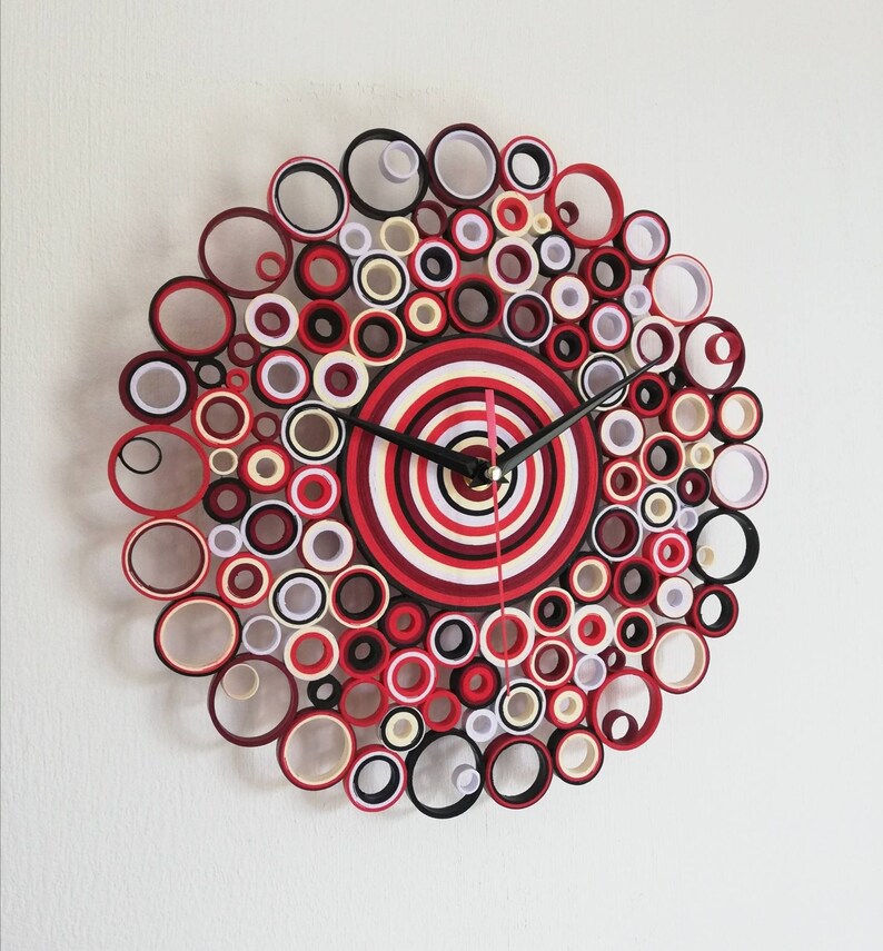 May include: A red, white, and black paper quilling clock with a circular design. The clock face is made up of concentric circles of red, white, and black paper.