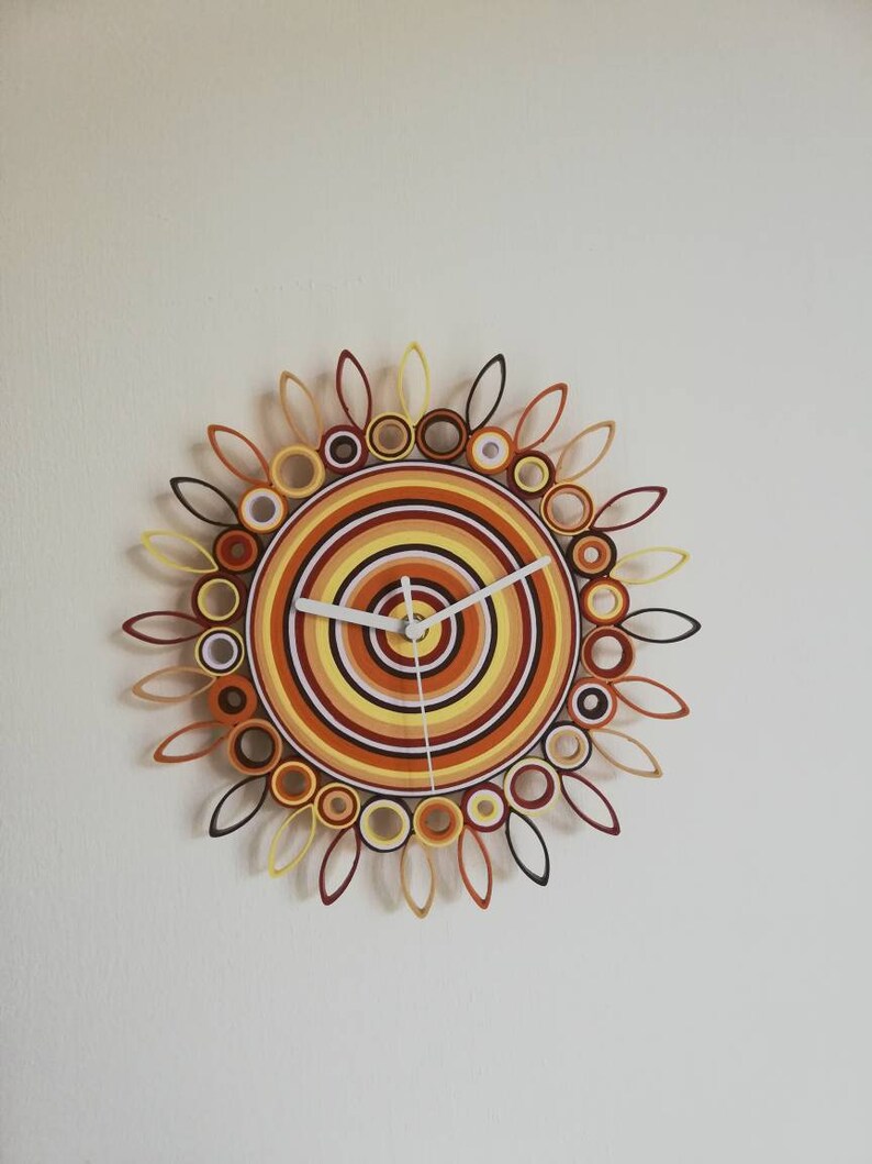 May include: A circular wall clock with a white face and black hands. The clock is made of paper and has a design of concentric circles in shades of orange, yellow, and brown. The outer edge of the clock has a design of paper leaves.