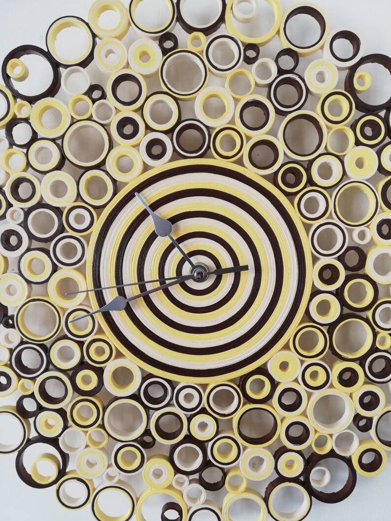 May include: A yellow and brown paper quilled clock with a concentric circle design. The clock face is surrounded by a ring of smaller circles.
