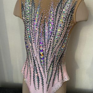 May include: A light pink leotard with a nude mesh overlay on the shoulders and upper back. The leotard is heavily embellished with iridescent rhinestones and crystals in a feather-like pattern. The design is symmetrical and covers the front and back.