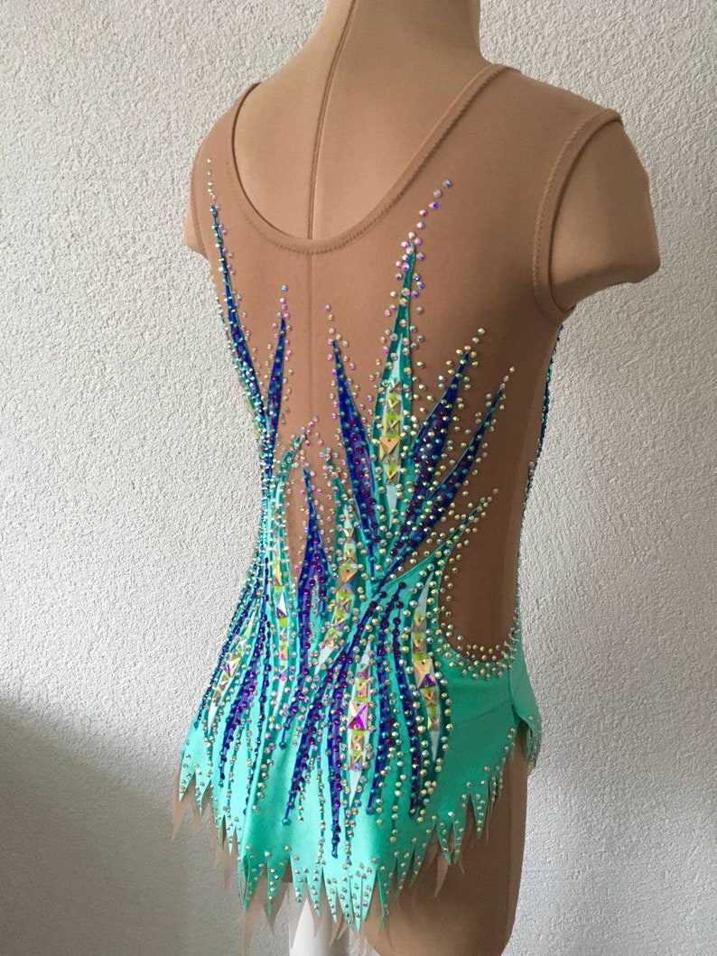 May include: A turquoise and blue leotard with a sheer back and a crystal design. The leotard has a scalloped hem and is decorated with rhinestones.