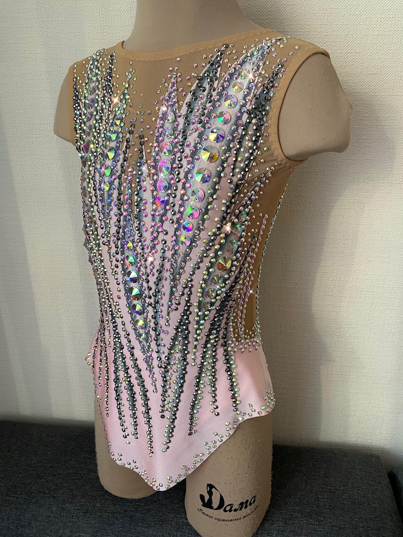 May include: A pink and nude leotard with a dazzling array of iridescent rhinestones and crystals. The sleeveless garment features a mesh overlay and a decorative pattern of sparkling embellishments, creating a glamorous look. The leotard is designed for gymnastics or dance performances.
