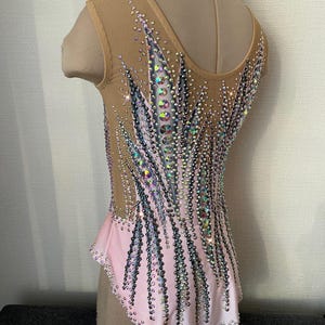 May include: A light pink and nude leotard with intricate crystal embellishments. The leotard features a V-neck and a mesh overlay on the upper body. The design includes vertical lines of crystals, creating a sparkling effect, perfect for gymnastics or dance.