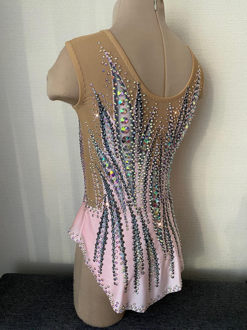 May include: A light pink and nude leotard with intricate crystal embellishments. The leotard features a V-neck and a mesh overlay on the upper body. The design includes vertical lines of crystals, creating a sparkling effect, perfect for gymnastics or dance.