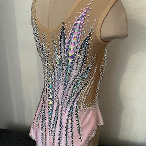 May include: A light pink and nude leotard with intricate rhinestone and sequin embellishments. The design features a gradient effect, with a concentration of sparkling details on the bodice and extending down the back. The leotard is sleeveless with a scoop neckline.