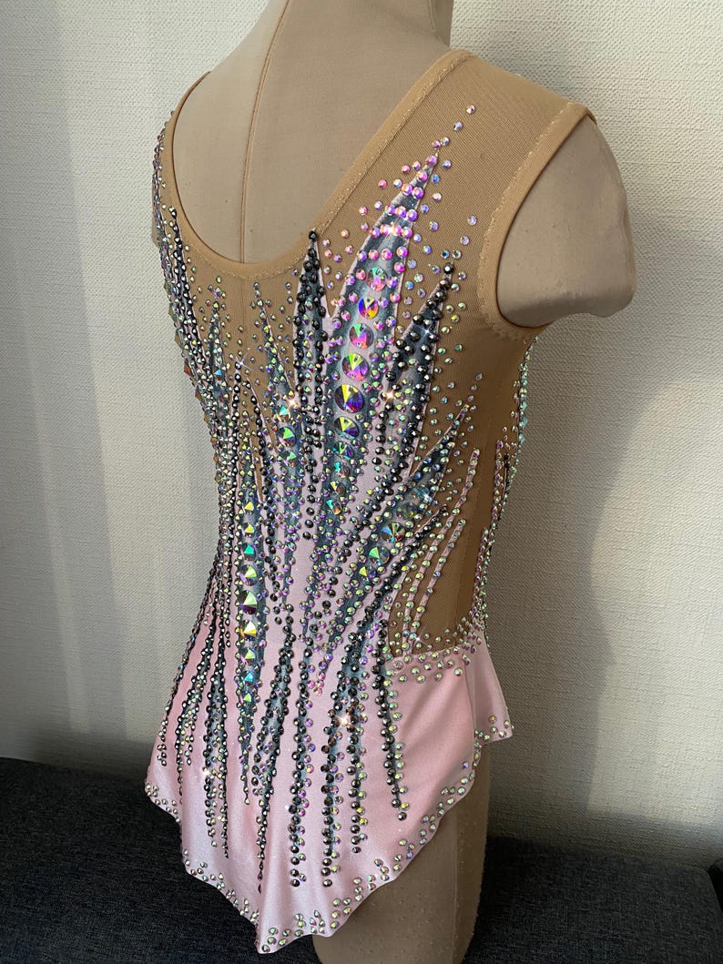 May include: A light pink and nude leotard with intricate rhinestone and sequin embellishments. The design features a gradient effect, with a concentration of sparkling details on the bodice and extending down the back. The leotard is sleeveless with a scoop neckline.
