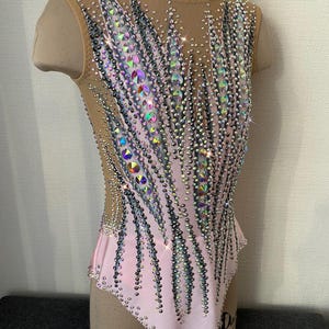 May include: A sleeveless pink leotard with a nude mesh overlay, heavily embellished with iridescent and silver rhinestones. The design features vertical patterns of rhinestones, creating a sparkling effect. The leotard is displayed on a mannequin.