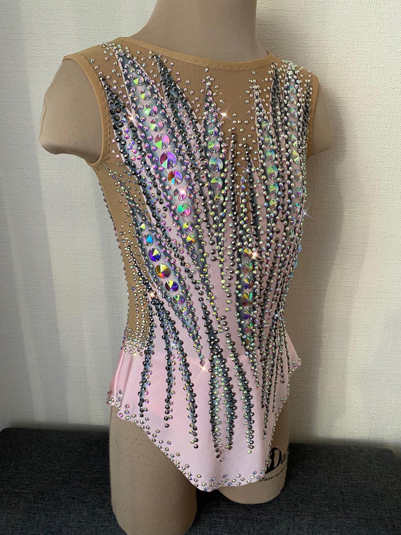 May include: A sleeveless pink leotard with a nude mesh overlay, heavily embellished with iridescent and silver rhinestones. The design features vertical patterns of rhinestones, creating a sparkling effect. The leotard is displayed on a mannequin.