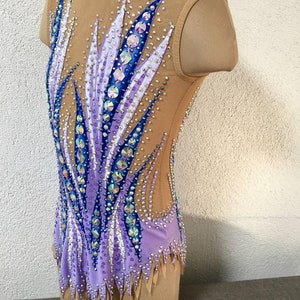 May include: A purple and blue leotard with a geometric design and rhinestones. The leotard has a scoop neck and sleeveless design. The leotard is on a dress form.